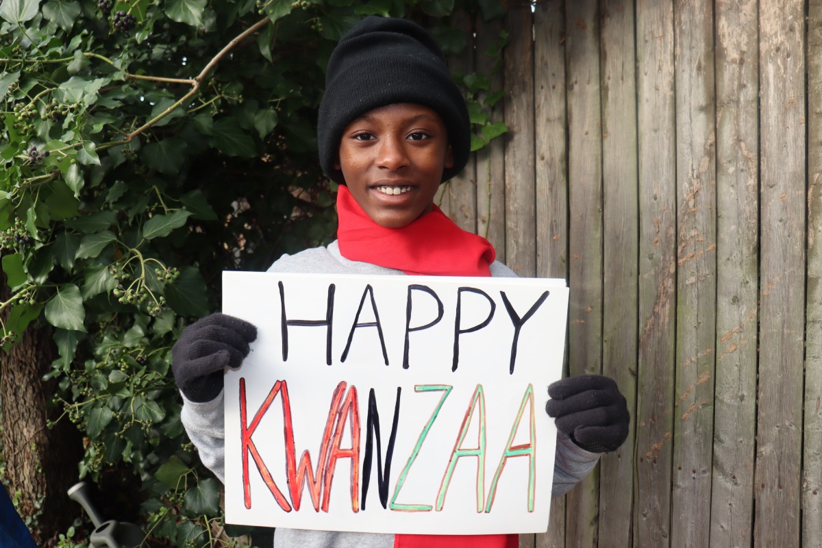 Kid in winter clothing holding Happy Kwanzaa sign outdoors-img