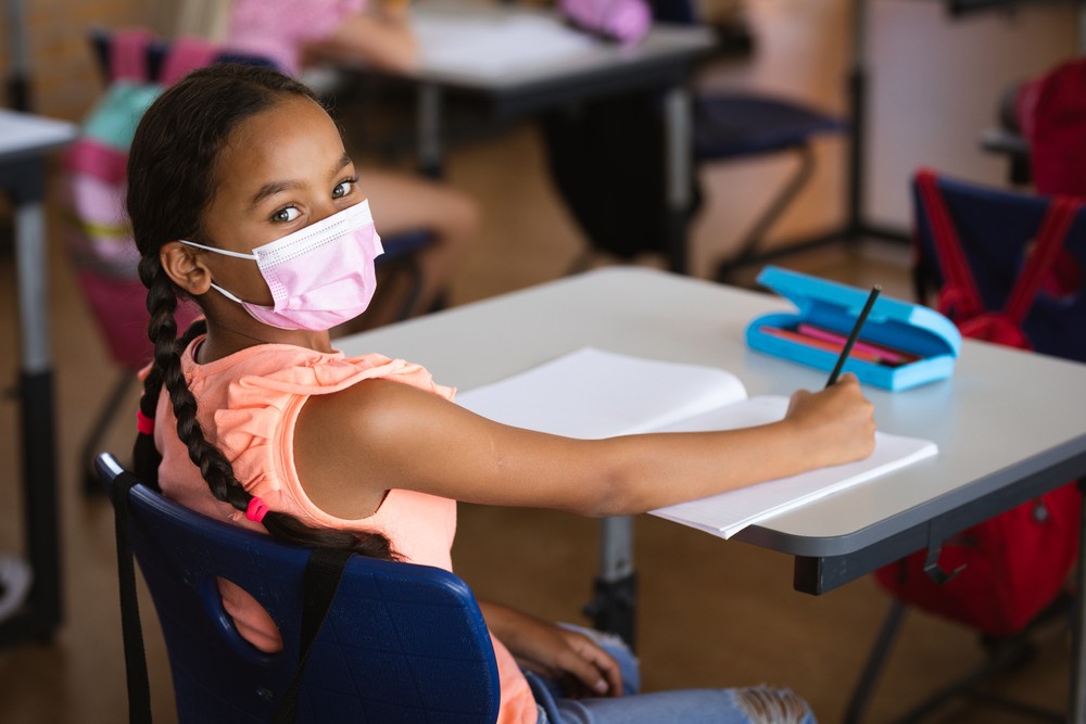 Portrait of african american girl wearing face mask sitting on her desk-img