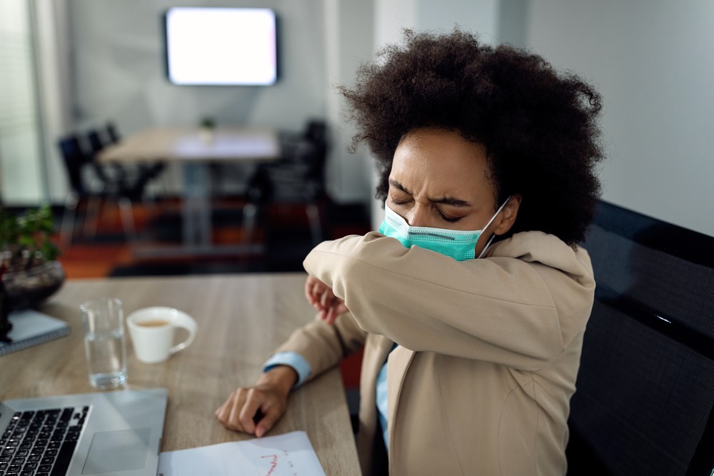 Black businesswoman coughing into elbow while wearing protective face mask-img